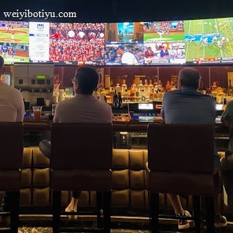 A group of people sit at a bar with their backs to the camera, watching multiple sports games displayed on large screens. The bar counter is lined with various bottles of liquor, and the atmosphere suggests a casual and relaxed social setting.