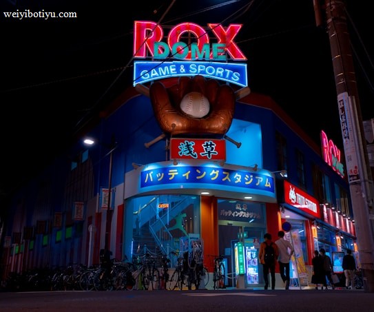 A vibrant, neon-lit facade of a gaming and sports establishment at night, featuring a large glove holding a baseball above the entrance. The building is adorned with colorful signs in both English and Japanese, with various advertisements and an illuminated sign that says 'ROX DOME'. Several people are walking towards the entrance, and bicycles are parked along the side.